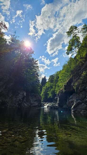A serene river flows between rocky cliffs and lush green trees under a bright sun and scattered clouds in a vibrant, clear blue sky. Peaceful nature scene.