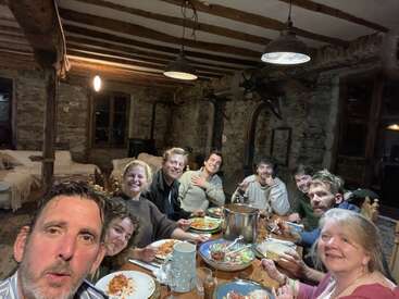 A group of nine people sit around a rustic wooden table, enjoying a meal together in a cozy, stone-walled room, smiling and sharing a joyful evening.