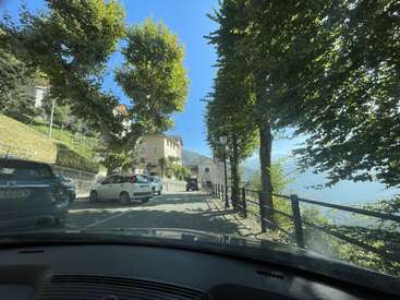 A narrow tree-lined road winds through a picturesque hillside village with parked cars, charming houses, and distant mountains under a bright blue, sunlit sky.