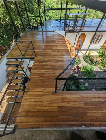 L'image représente un escalier en colimaçon avec des rampes noires et des marches en bois, situé dans un bâtiment moderne avec un mur en verre et un jardin luxuriant à l'extérieur.