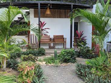 A imagem mostra um jardim tropical sereno com vegetação exuberante e plantas vibrantes, levando a uma área de estar externa aconchegante com dois bancos de madeira.