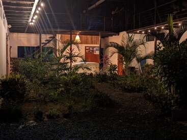 A imagem retrata um pátio sereno à noite, com vegetação exuberante, um edifício branco e uma luminária quente, exalando um ambiente tranquilo.