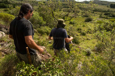 Two men are hiking through a lush, green landscape. One carries a machete on his belt. They walk among trees and rocks, exploring nature together.