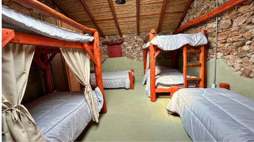 This rustic dormitory features wooden bunk beds and single beds with gray bedding, stone walls, a sloped wooden ceiling, cozy natural lighting, and a tranquil atmosphere.