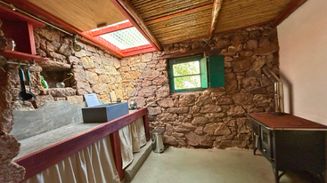 This rustic kitchen features stone walls, a wood stove, natural light from a skylight, a small window, concrete sink, and wooden shelving with minimal decor.