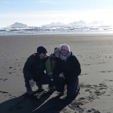 Three people bundled in winter clothing kneel and pose on a dark sandy beach with crashing ocean waves and rocky islands visible in the distant background.