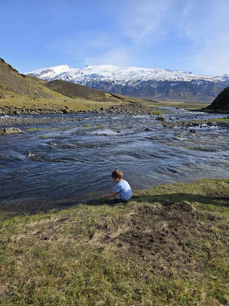 A child sits by a river, surrounded by grassy banks. Snow-capped mountains rise in the distance under a clear blue sky, creating a serene scene.