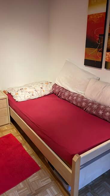 A tidy bedroom features a single bed with red bedding, patterned pillows, a wooden nightstand, a matching red rug, and colorful abstract art on the wall.