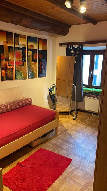 This cozy bedroom features a single bed with red bedding, modern artwork, wooden furniture, a full-length mirror, a red rug, and a window with dark curtains.