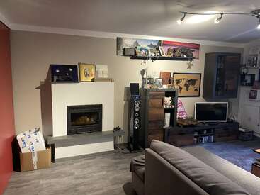 A cozy living room with a fireplace, TV, modern shelves, world map decor, boxes in the corner, speakers, a gray sofa, and various decorative items.