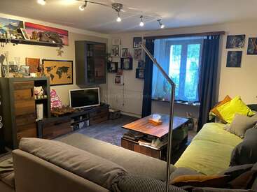 A cozy living room with a large sofa, wooden furniture, a coffee table, wall art, shelves, TV, window with curtains, books, and decorative items.