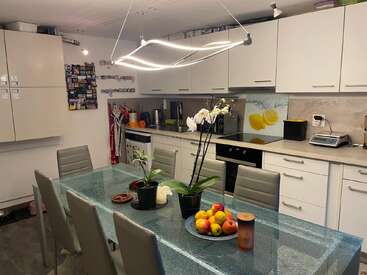 A modern kitchen with white cabinets, a glass dining table, gray chairs, fruit bowl, orchids, unique light fixture, magnets on the wall, and various appliances.