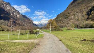 A peaceful country road winds through green fields, flanked by tall mountains. The sky is blue with clouds, and snowy peaks are visible in the distance.