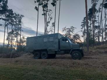 Un grand camion militaire vert à six roues est garé sur un terrain herbeux, entouré de grands arbres fins, sous un ciel nuageux au crépuscule.