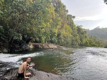 A man sits smiling at the riverbank, giving a thumbs up. People swim in the natural pool beside lush, green forest under a bright sky.