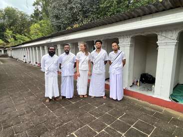 Five people dressed in white traditional attire stand together outdoors on a paved walkway, with white pillars and greenery in the background, looking peaceful and united.