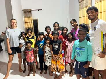 A cheerful group of children and a few adults pose together indoors, smiling. The room has yellow curtains, white walls, and a bright, welcoming atmosphere.