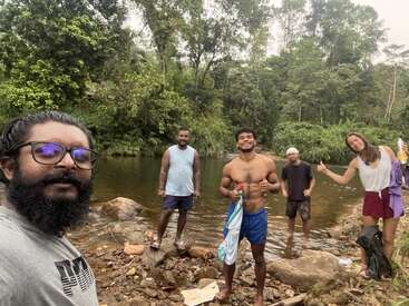 Five friends stand by a river, surrounded by lush greenery. They smile and pose for a group selfie, enjoying the outdoors and nature together.