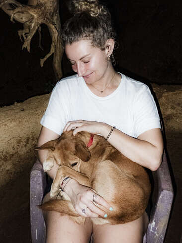 Une femme est assise dans un fauteuil, souriant doucement tout en berçant un chien brun lové sur ses genoux. La scène rayonne de chaleur, de confort et d'affection.