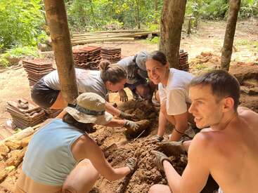 Five people work together outdoors, kneeling on the ground, handling soil with gloved hands. Surrounded by trees, stacked tiles, and nature, they appear happy and collaborative.