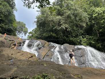 Four people explore a beautiful, rocky waterfall surrounded by lush green forest. Water cascades down the smooth rocks as one person stands in the foreground.
