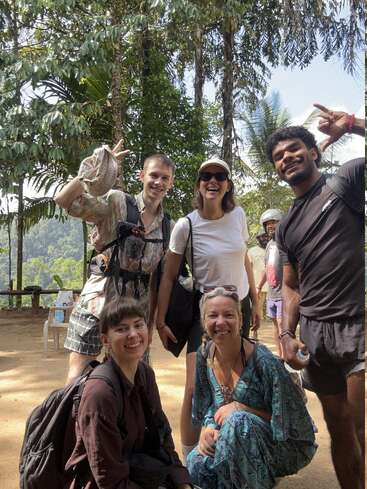 Five friends pose cheerfully outdoors amidst lush greenery, smiling under the sunlight. Two kneel in front, while three stand behind, making playful hand gestures.