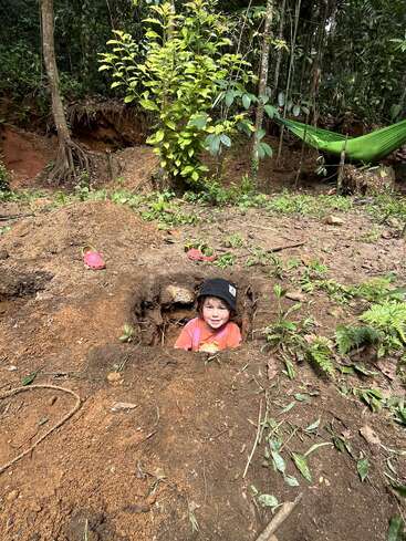 A young child wearing a black hat and red shirt sits in a freshly dug hole in a forest, surrounded by dirt, greenery, and a green hammock.