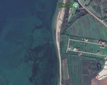 Dieses Satellitenbild zeigt ein Küstengebiet in der Nähe von Actium in Griechenland mit einem schmalen Strand, klarem blauem Wasser, grünen Feldern, vereinzelten Gebäuden und einem kleinen Straßennetz.