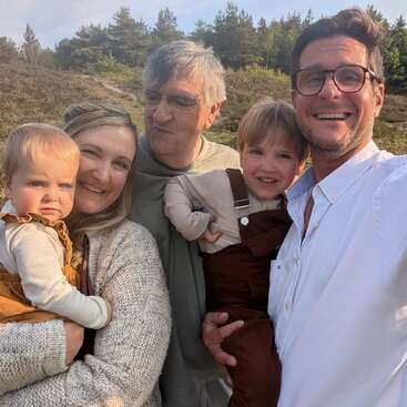 Eine glückliche fünfköpfige Familie posiert im Freien und lächelt herzlich. Zwei kleine Kinder werden von Erwachsenen im Arm gehalten, umgeben von viel Grün und unter einem sonnigen, klaren Himmel. Ein fröhlicher Moment, festgehalten.