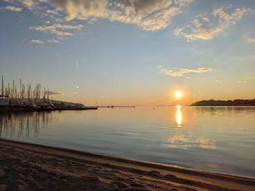 Eine ruhige Strandszene bei Sonnenuntergang, mit der Sonne, die sich auf dem sanften Wasser spiegelt, Booten, die links am Ufer liegen, weichem Sand und vereinzelten Wolken am friedlichen Himmel.