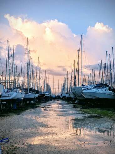 Bei Sonnenuntergang reihen sich Segelboote zu beiden Seiten eines nassen, spiegelnden Weges aneinander. Sanfte Wolken und blauer Himmel schaffen eine friedliche Atmosphäre am Yachthafen.