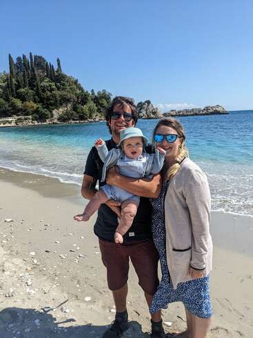 Eine glückliche Familie steht an einem Sandstrand am Meer und hält ein Baby im Arm. Die Sonne scheint, und im Hintergrund sind Bäume und Felsen zu sehen.