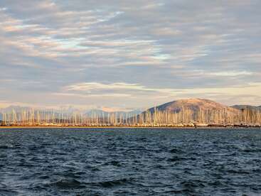 Eine ruhige Meereslandschaft mit dunklem, plätscherndem Wasser im Vordergrund. Zahlreiche Segelbootsmasten säumen das Ufer. Sanfte Hügel und ein bewölkter Himmel runden das Bild ab.