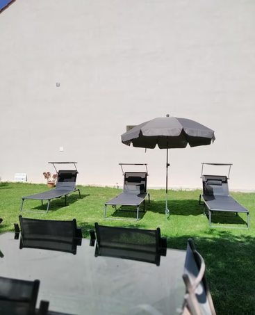 Auf diesem Bild sind vier Liegestühle und ein großer Sonnenschirm auf grünem Rasen zu sehen, im Vordergrund stehen ein Glastisch und vier Stühle vor einer schlichten Wand.
