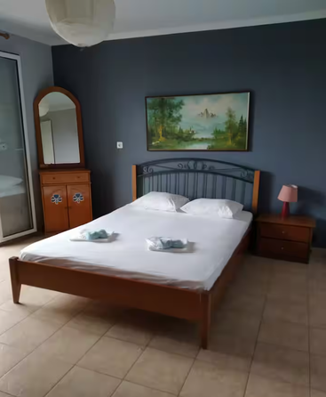 Ein gemütliches Schlafzimmer mit einem Holzbett, zwei Kissen, gefalteten Handtüchern, einer Nachttischlampe, einer Kommode mit Spiegel, einem Landschaftsgemälde, Fliesenboden und einer blauen Akzentwand.