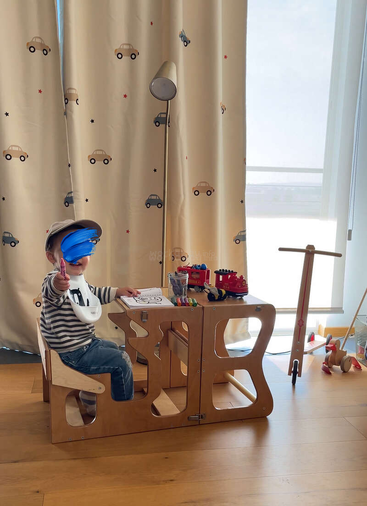 Ein kleines Kind sitzt an einem Holztisch mit Malutensilien und zeigt auf einen Marker. Der Raum hat verspielte Vorhänge, Spielzeug und viel natürliches Licht.