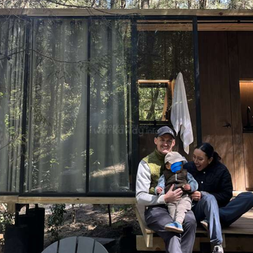 A family of three sits happily on the porch steps of a modern cabin in the woods, surrounded by trees and nature, enjoying a peaceful moment together.