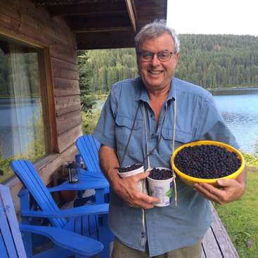 The image depicts a man standing on a porch, holding two containers of blueberries and a yellow bowl, with a serene lake and forested hillside visible in the background.