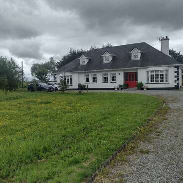 A white house with a dark roof and red door sits amid a grassy lawn, yellow flowers, gravel driveway, two cars, cloudy sky, and surrounding trees.