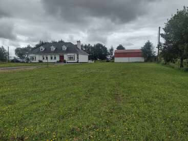 This image shows a white house with a red door and nearby barn, surrounded by green grass and trees under a cloudy, overcast sky. Peaceful countryside scene.