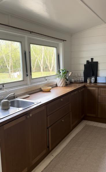 Das Bild zeigt eine Küche mit Spüle, Herd und Kühlschrank, mit weißen Schränken und Arbeitsflächen, mit einem Fenster über der Spüle und einer Tür auf der rechten Seite.