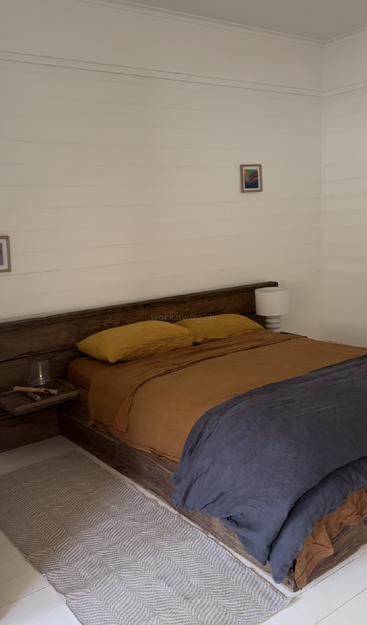 Das Bild zeigt ein gemütliches Schlafzimmer mit einem hölzernen Bettgestell, einem weißen Nachttisch und einem grauen Teppich, auf dem eine braune Bettdecke und zwei Kissen liegen.