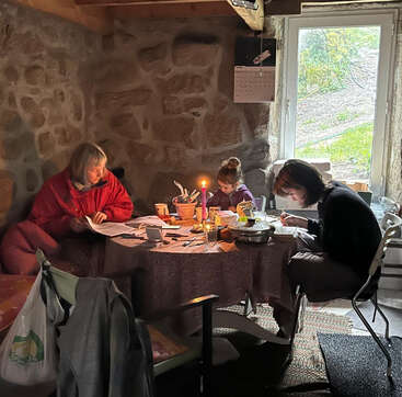 Three people sit around a table in a rustic stone room, illuminated by a candle, concentrating on reading, writing, or drawing amidst various household items.