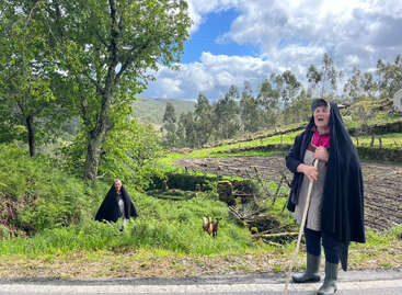 Two women wearing black cloaks stand in a lush, green rural landscape with trees, goats, and blue sky, creating a serene, pastoral, countryside scene.