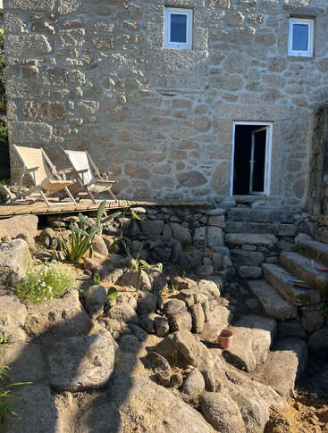 A rustic stone house with three windows, an open door, two deck chairs on a wooden platform, rocky steps, and some scattered plants basking in sunlight.