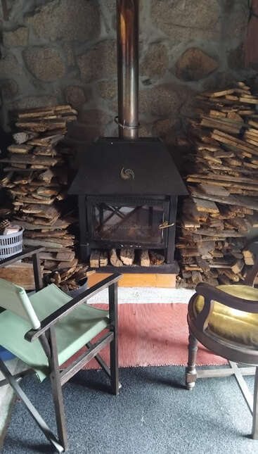 A cozy room features a wood stove with stacked firewood on both sides, two chairs, a stone wall backdrop, and a red rug on the floor.