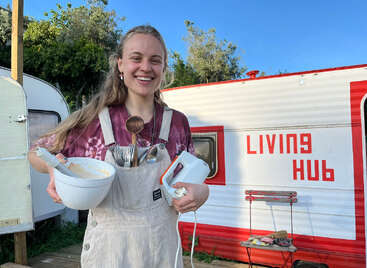 A smiling woman stands outdoors by a trailer labeled "LIVING HUB," holding a mixing bowl, utensils, and hand mixer, wearing an apron and a tie-dye t-shirt.
