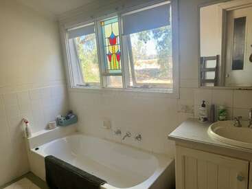 Ein helles Badezimmer mit einer weißen Badewanne, gefliesten Wänden und einem einfachen Waschbecken. Große Fenster, eines davon mit Buntglas, bieten einen Blick auf das Grün draußen und sorgen für Farbe und natürliches Licht.