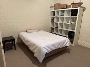 Das schlichte Schlafzimmer besteht aus einem Einzelbett mit weißer Bettwäsche, einem Beistelltisch und einem weißen, würfelförmigen Regal, in dem Bücher, Kisten und andere Dinge untergebracht sind.