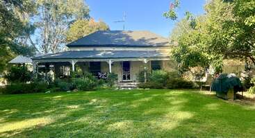 Ein charmantes Landhaus mit einer breiten Veranda, umgeben von üppigen Bäumen und Grün. Der gepflegte Rasen und die verstreuten Gartenbeete schaffen eine friedliche Atmosphäre.